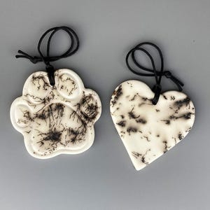 May include: Two ceramic ornaments, one shaped like a paw print and the other a heart, with black and white swirling patterns. Both ornaments have black cords for hanging.