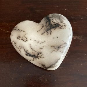 May include: A heart-shaped ornament with a white surface, adorned with delicate black, vein-like patterns. The object rests on a dark wooden surface. The design suggests a unique, handcrafted item, possibly ceramic or stone.