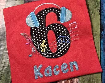 May include: A red t-shirt featuring an embroidered black number six with white polka dots, topped with light blue headphones. The design includes musical notes and a staff, with the name Kacen embroidered in light blue letters below the number.