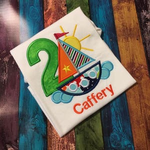 May include: A white t-shirt with an embroidered design of a sailboat, a sun, and the number 2. The name "Caffery" is embroidered below the design.