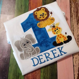 May include: White t-shirt with a blue number 1 and the name "DEREK" embroidered in blue. The shirt features embroidered cartoon animals: a lion, a giraffe, and an elephant.