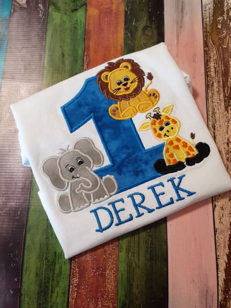 May include: White t-shirt with a blue number 1 and the name "DEREK" embroidered in blue. The shirt features embroidered cartoon animals: a lion, a giraffe, and an elephant.