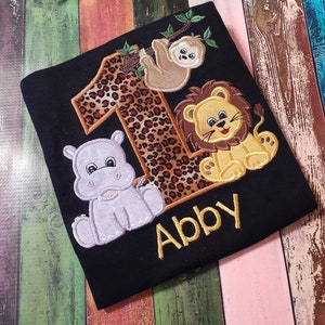 May include: Black t-shirt with embroidered animal and number designs. Features a leopard print number 1, a sloth, a lion, and a hippo. The name "Abby" is embroidered in gold below the animals. A fun, colorful design.