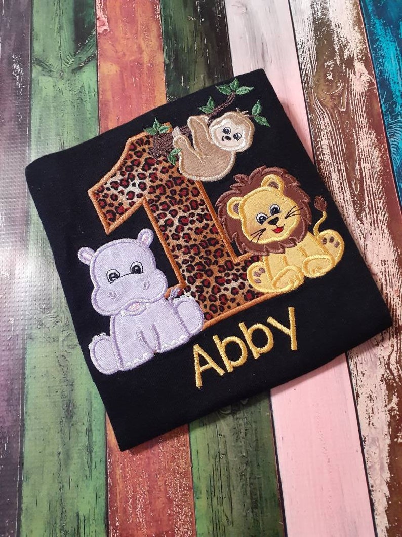 May include: Black t-shirt with embroidered animal and number designs. Features a leopard print number 1, a sloth, a lion, and a hippo. The name "Abby" is embroidered in gold below the animals. A fun, colorful design.