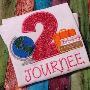 May include: A white t-shirt with a pink glittery number 2, a globe, and three suitcases. The text "JOURNEE" is embroidered in pink below the number.