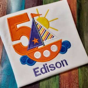 May include: A white t-shirt with an orange number 5, a blue and orange sailboat with a yellow sun, and the name Edison embroidered in purple.