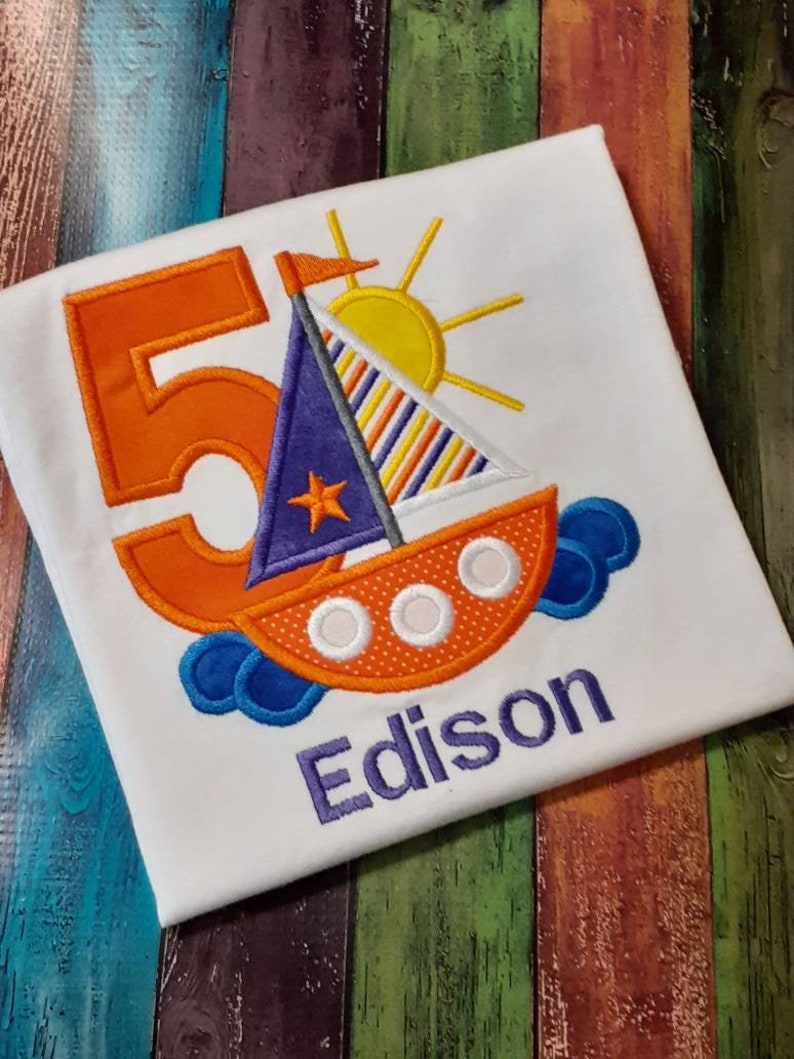 May include: A white t-shirt with an orange number 5, a blue and orange sailboat with a yellow sun, and the name Edison embroidered in purple.
