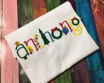 May include: White t-shirt embroidered with the name "Anthony" in colorful Christmas-themed lettering. The letters are decorated with festive elements like a Christmas tree, lights, ornaments, and a wreath. The shirt is folded on a multicolored wooden surface.