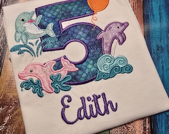 May include: A white t-shirt featuring an embroidered design of a large number five with a blue scale pattern, surrounded by three dolphins in light blue, pink, and purple, an orange balloon, teal waves, and the name Edith in purple script.