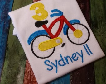 May include: White t-shirt with a red, blue, and yellow bicycle design, the number 3, and the name "Sydney" embroidered in blue.