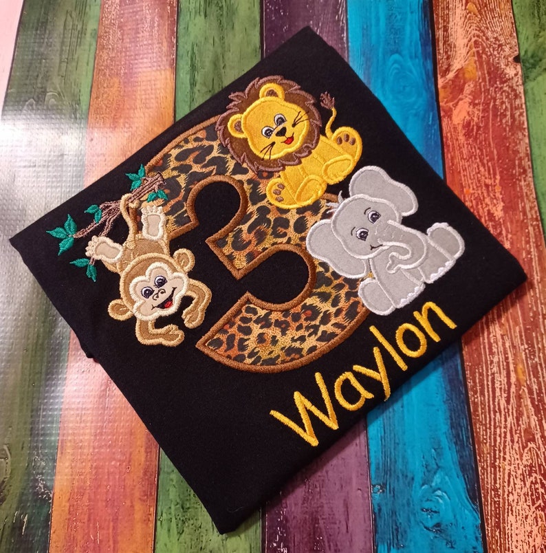 May include: Black t-shirt with embroidered jungle animals and the number 2 in a leopard print design. Features a monkey, lion, and elephant. The name "Waylon" is embroidered in yellow.