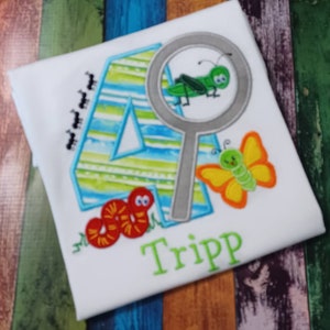 May include: A white t-shirt with a blue and green number four, a magnifying glass with a green grasshopper inside, a yellow butterfly, a red worm, and the name "Tripp" embroidered on the front.