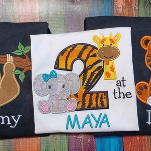 May include: Three embroidered shirts with animal designs. One shirt has a sloth hanging from a branch with "Mommy" below. Another has a number 2 with a giraffe and elephant, and the name "MAYA". The third shirt has a tiger with "Daddy".