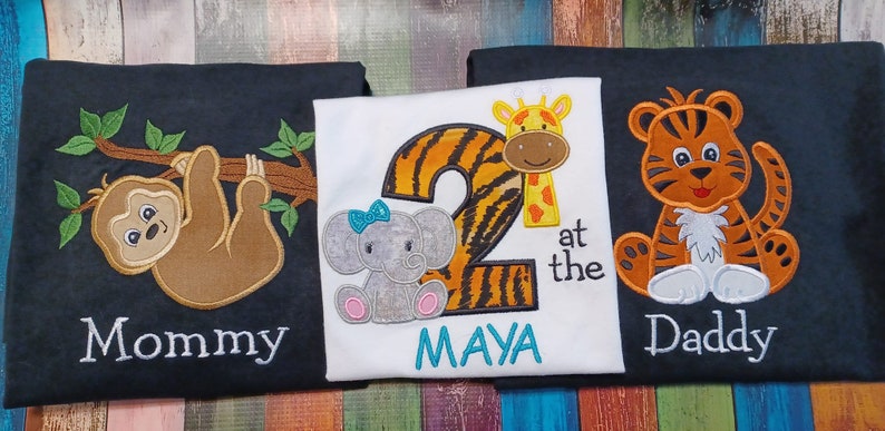 May include: Three embroidered shirts with animal designs. One shirt has a sloth hanging from a branch with "Mommy" below. Another has a number 2 with a giraffe and elephant, and the name "MAYA". The third shirt has a tiger with "Daddy".