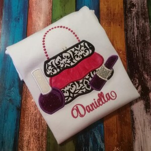 May include: White t-shirt with an embroidered design of a black and white purse with a pink handle, a pink lipstick, and a purple nail polish bottle. The name "Daniella" is embroidered in pink below the design.