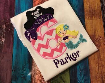 May include: A white t-shirt with a pink number six with a chevron pattern, a purple pirate hat with a skull and crossbones, and a mermaid with a turquoise tail and a yellow flower in her hair. The name "Parker" is embroidered in purple below the design.