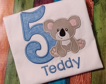 Könnte beinhalten: Ein weißes T-Shirt mit einer blauen Zahl 5 und einem grauen Koalabär, der darauf gestickt ist. Der Name "Teddy" ist unter dem Koalabär gestickt.