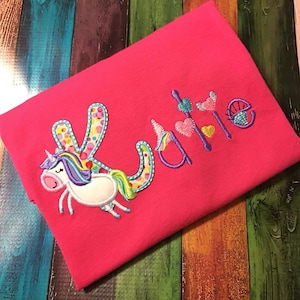 May include: Pink t-shirt with a unicorn and the name "Katie" embroidered on it. The unicorn is white with a rainbow mane and tail. The letters are colorful and have a polka dot pattern.