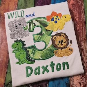 May include: White t-shirt with embroidered design featuring the text "WILD and 3" and the name "Daxton". The design includes an elephant, a dinosaur, a lion, and a yellow dinosaur. The number 3 is green with a leaf pattern.