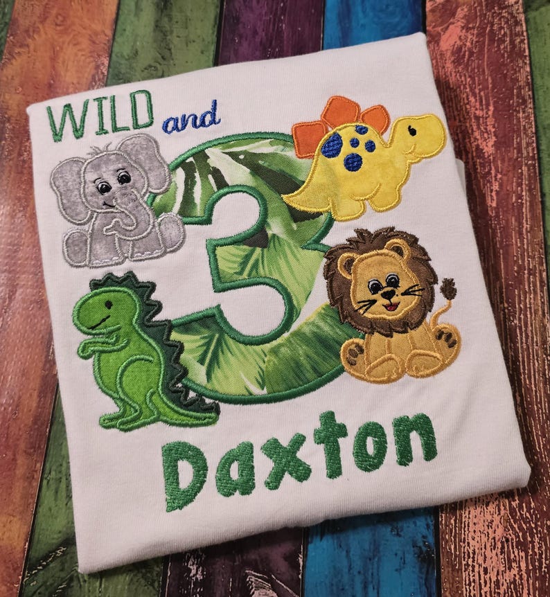May include: White t-shirt with embroidered design featuring the text "WILD and 3" and the name "Daxton". The design includes an elephant, a dinosaur, a lion, and a yellow dinosaur. The number 3 is green with a leaf pattern.