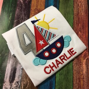 May include: A white t-shirt with a gray number 4, a red, blue, and white sailboat with a yellow sun, and the name "CHARLIE" in red embroidery.