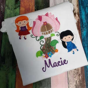 May include: A white t-shirt with an embroidered design of two fairies and a pink flower house. The name "Macie" is embroidered in purple below the design.
