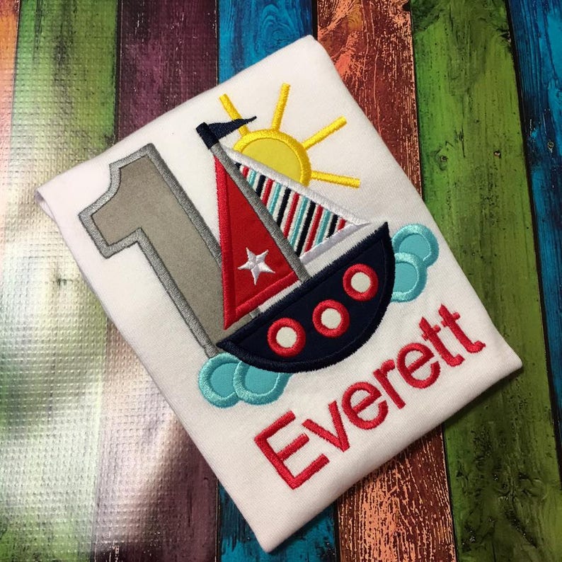 May include: A white onesie with a red, white, and blue sailboat, a yellow sun, and the number one in gray. The name Everett is embroidered in red below the sailboat.