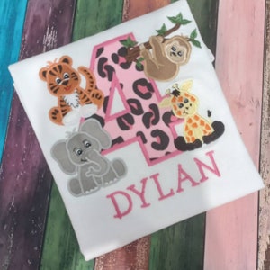 May include: White t-shirt with a pink number 4 featuring a leopard print design. The shirt is decorated with embroidered cartoon animals, including a tiger, elephant, giraffe, and sloth. The name "DYLAN" is embroidered in pink.
