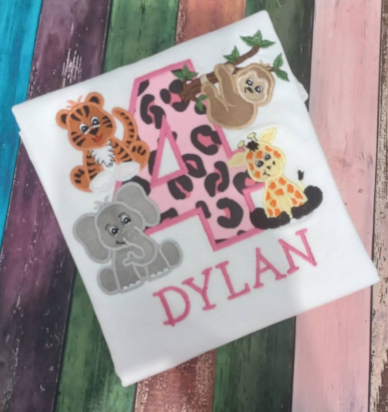 May include: White t-shirt with a pink number 4 featuring a leopard print design. The shirt is decorated with embroidered cartoon animals, including a tiger, elephant, giraffe, and sloth. The name "DYLAN" is embroidered in pink.