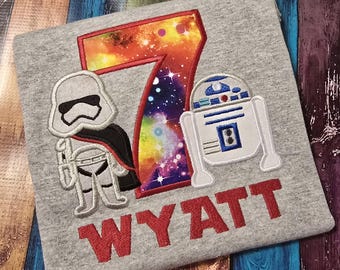 May include: A heather gray t-shirt featuring an embroidered design of a large number 7 with a colorful galaxy print, flanked by a white stormtrooper and a white R2-D2 robot. The name WYATT is embroidered in red capital letters below the design.