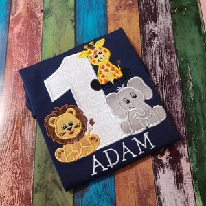 May include: Navy blue shirt with embroidered safari animals and the number 1. Features a giraffe, lion, and elephant. The name "ADAM" is embroidered below the animals. A cute and colorful design for a child's birthday.