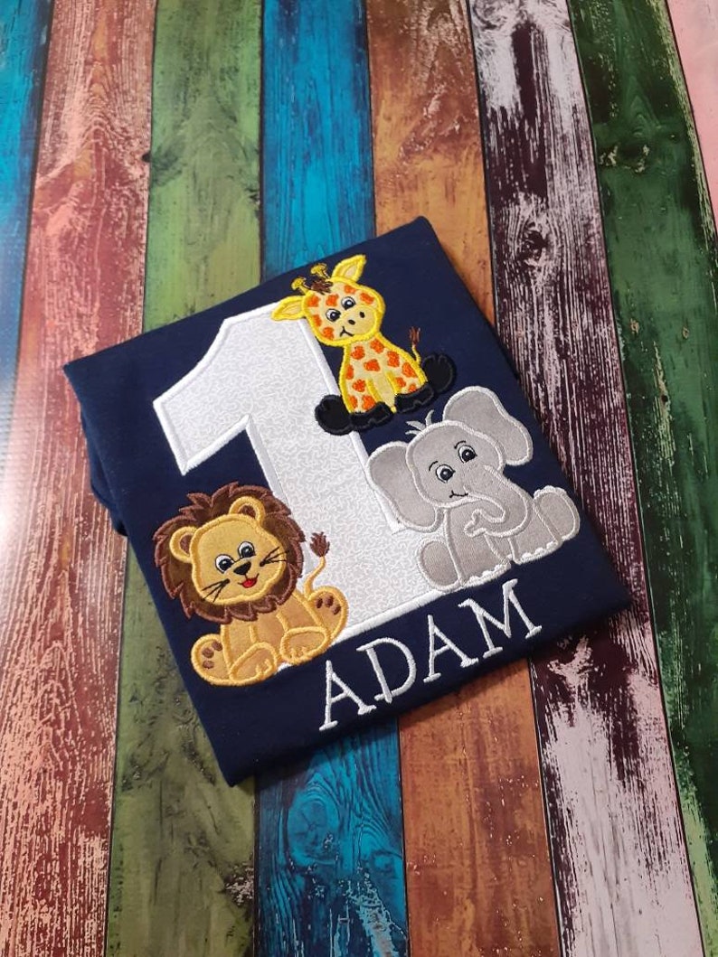 May include: Navy blue shirt with embroidered safari animals and the number 1. Features a giraffe, lion, and elephant. The name "ADAM" is embroidered below the animals. A cute and colorful design for a child's birthday.