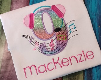 May include: A white t-shirt with a pink, blue, and green glittery number nine with headphones and musical notes. The name "mackenzie" is embroidered in pink below the number.