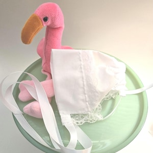 May include: A pink flamingo plush toy with an orange beak sits on a green plate with a white satin ribbon and a white lace trimmed bib.