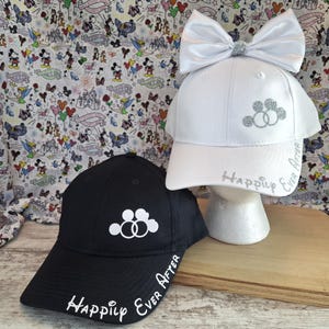 May include: Two baseball caps, one black and one white, with the words "Happily Ever After" and a double circle design. The white cap has a large white bow with a silver glitter centre. The caps are on a wooden surface.