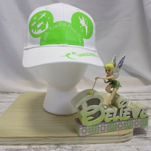 May include: White baseball cap with a green Mickey Mouse ear design. The design includes the text "Faith, Trust & Pixie Dust" and a silhouette of Tinkerbell. The cap is displayed on a white mannequin head with a wooden base. A Tinkerbell figurine is on the base with a "Believe" sign.
