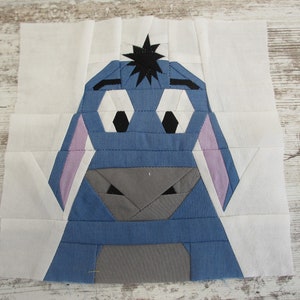 May include: A blue and gray fabric patchwork quilt block featuring a cartoon character with a star on its head. The character has large eyes and a small nose.