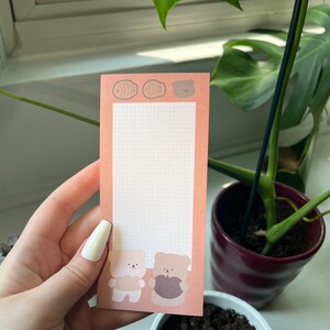 May include: A rectangular peach-colored notepad featuring a white grid pattern in the center. The top border displays two fish-shaped icons and a small bear head. The bottom section shows two cartoon bears, one light and one dark, standing side by side.