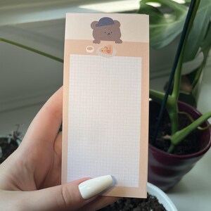 May include: A rectangular notepad with a light peach background featuring a grid pattern for writing. At the top, a brown cartoon bear wearing a blue hat is illustrated above a small plate with a slice of pizza and a small bowl of dipping sauce.