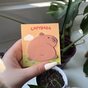 May include: A square sticky note featuring a brown capybara illustration on a light orange background with a green grassy field, a white cloud, and the text CAPYBARA printed in orange at the top. The note has a die-cut shape of a capybara.