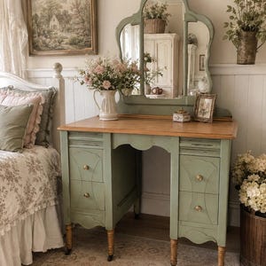 May include: A sage green vanity table with a natural wood top, featuring two columns of two drawers each with small gold knobs. The vanity includes a tall, arched mirror attached to the back, supported by four turned wooden legs with casters.