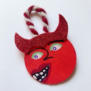 May include: Red felt ornament shaped like a devil's face with glitter horns, green and purple eyes, and a painted smile.