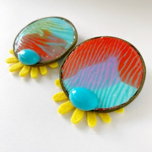 May include: A pair of colorful earrings with a blue and red abstract design, set in a gold-toned metal frame. Each earring is attached to a yellow felt flower.