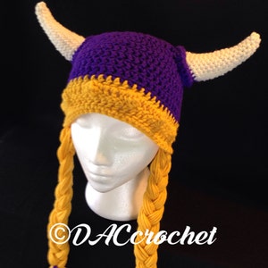 Pu&ograve; includere: Cappello vichingo all'uncinetto viola e oro con corna bianche e trecce gialle. Il cappello &egrave; su una testa di manichino bianca. Il testo "DACcrochet" &egrave; sul cappello.