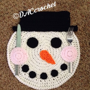 May include: White crocheted snowman placemat with black eyes, a carrot nose, and pink cheeks. The placemat has a black crocheted hat and a fork and knife are placed on the sides.