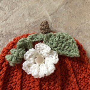 Op de afbeelding: Een gebreide pompoenmuts met een witte bloem en groene bladeren erop. De muts is oranje en heeft een textuur.