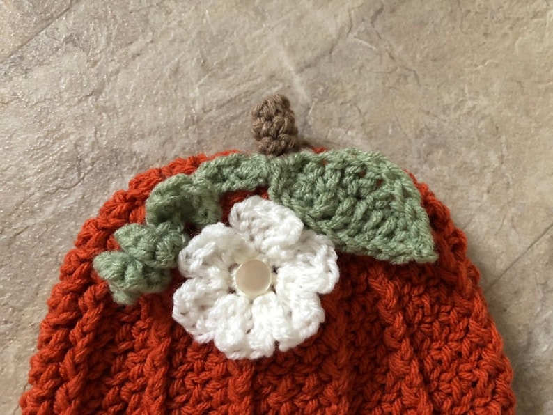 Op de afbeelding: Een gebreide pompoenmuts met een witte bloem en groene bladeren erop. De muts is oranje en heeft een textuur.