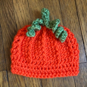 May include: An orange crochet hat shaped like a pumpkin, with green crochet leaves and stem on top. The hat is made of a textured, knitted material. Perfect for Halloween or fall-themed events.