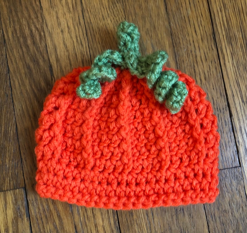 May include: An orange crochet hat shaped like a pumpkin, with green crochet leaves and stem on top. The hat is made of a textured, knitted material. Perfect for Halloween or fall-themed events.