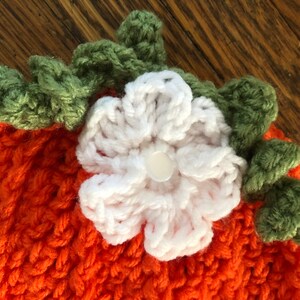May include: Close-up of a handmade orange crochet pumpkin hat with a white flower and green leaves. The hat is made of a textured yarn and features a button in the center of the flower. Perfect for fall and Halloween.